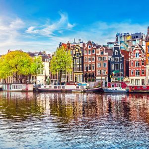 Güneşli gökyüzü altında Amsterdam kanalına paralel sıralanmış ikonik renkli evleri, kanal teknelerini ve sudaki yansımaları gösteren yüksek çözünürlüklü şehir duvar kağıdı görseli.