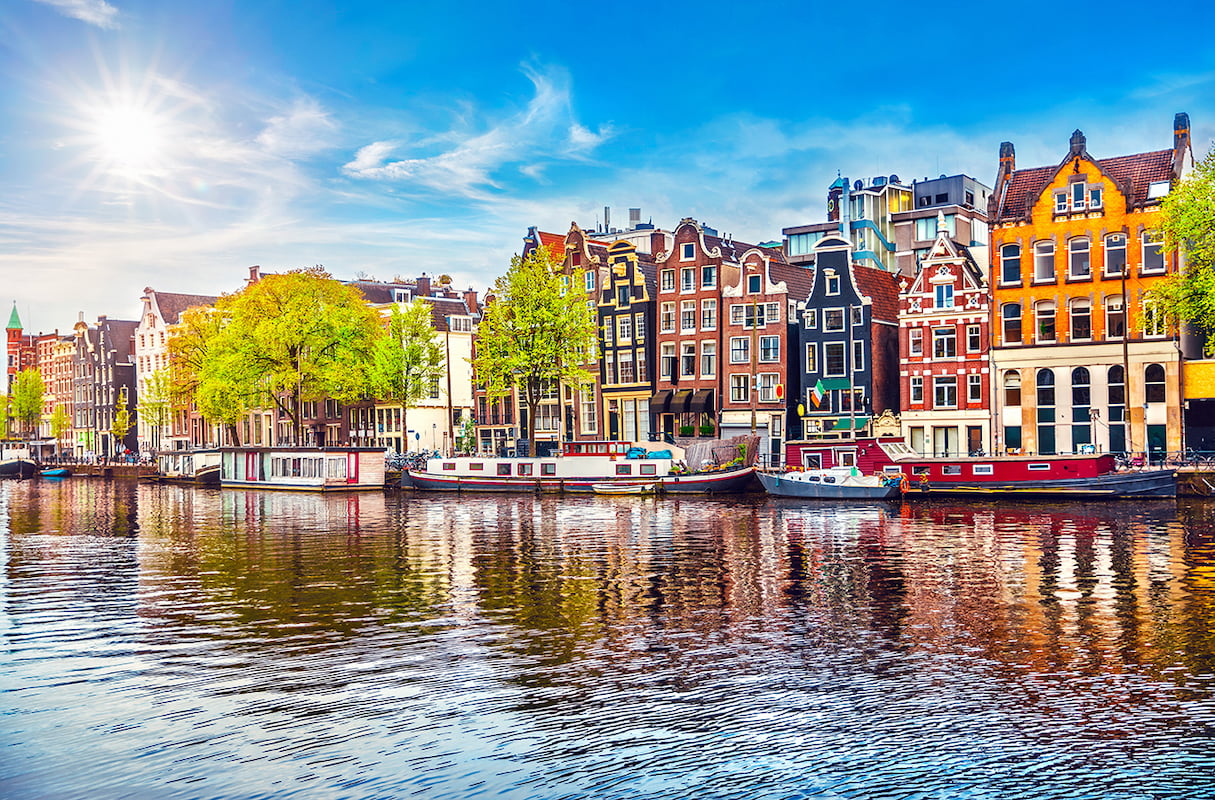 Güneşli gökyüzü altında Amsterdam kanalına paralel sıralanmış ikonik renkli evleri, kanal teknelerini ve sudaki yansımaları gösteren yüksek çözünürlüklü şehir duvar kağıdı görseli.