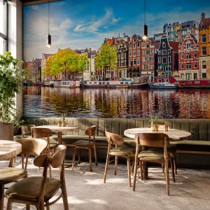 Amsterdam kanalı kıyısındaki renkli tarihi evler ve su üzerindeki yansımalar manzarası; ahşap kafe masaları, deri sandalyeler ve dikey sarkıt lambalarla restoran ile kafelerde canlı bir Avrupa şehir atmosferi yaratır.