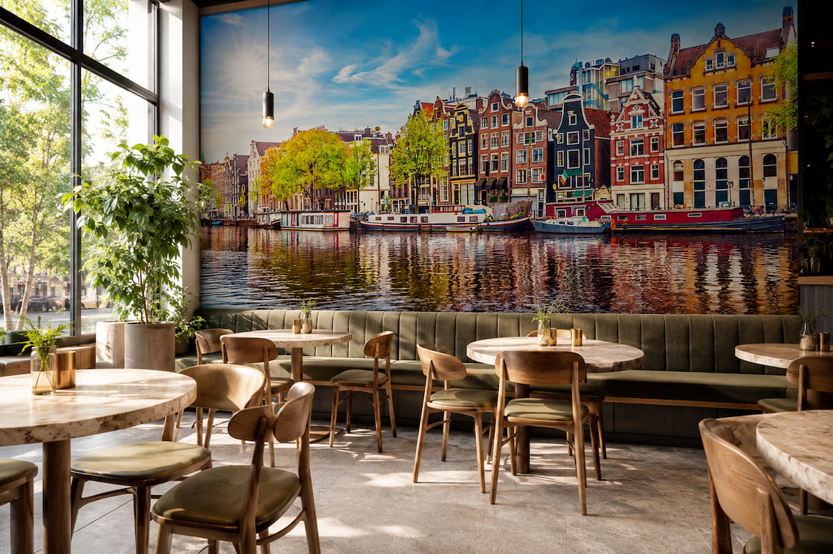 Amsterdam kanalı kıyısındaki renkli tarihi evler ve su üzerindeki yansımalar manzarası; ahşap kafe masaları, deri sandalyeler ve dikey sarkıt lambalarla restoran ile kafelerde canlı bir Avrupa şehir atmosferi yaratır.