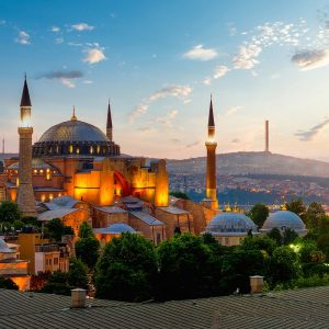 Gün batımının altın ve mavi tonlu gökyüzü altında ışıklandırılmış tarihi Ayasofya Camii'ni, minarelerini, kubbelerini ve arka plandaki İstanbul şehir silüetini gösteren yüksek çözünürlüklü duvar kağıdı görseli.