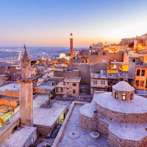 Alacakaranlıkta ışıkları yanan tarihi Mardin taş evlerini, minareleri, tarihi kubbeleri ve sonsuz Mezopotamya ovasını gösteren yüksek çözünürlüklü panoramik duvar kağıdı görseli.