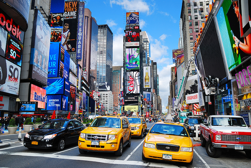 Ön planda akan trafikteki ikonik sarı taksiler, arkada Times Square'in dev dijital reklam ekranları, yüksek gökdelenler ve mavi gökyüzünü gösteren yüksek çözünürlüklü şehir duvar kağıdı görseli.