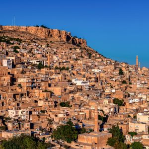 Berrak mavi gökyüzü altında dağ yamacına kurulmuş tarihi Mardin taş evlerini, cami minarelerini ve Mardin Kalesi'ni gösteren yüksek çözünürlüklü panoramik şehir duvar kağıdı görseli.