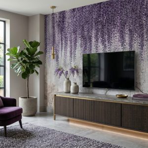 Eskitme mermer ve sıva dokulu açık gri zemin üzerine tavandan aşağı süzülen mor mor salkım (wisteria) çiçekleri temalı duvar kağıdı; ahşap TV konsolu, televizyon, yeşil saksı bitkisi ve mor berjerle salona canlı, modern ve şık bir hava katar.
