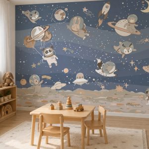 Gezegenler, yıldızlar ve astronot kıyafetli sevimli hayvan illüstrasyonlu duvar kağıdının uygulandığı; ahşap çocuk masası, ahşap oyuncak kulesi ve desenli halıyla döşenmiş oyun odası görünümü.