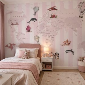 Pastel pembe ve beyaz dikey çizgili fon üzerinde dünya haritası, sıcak hava balonları, uçaklar ve sevimli deniz/kara hayvanları illüstrasyonlu çocuk odası duvar kağıdı; pembe yatak, komodin, abajur ve halıyla zarif bir mekan kurgular.