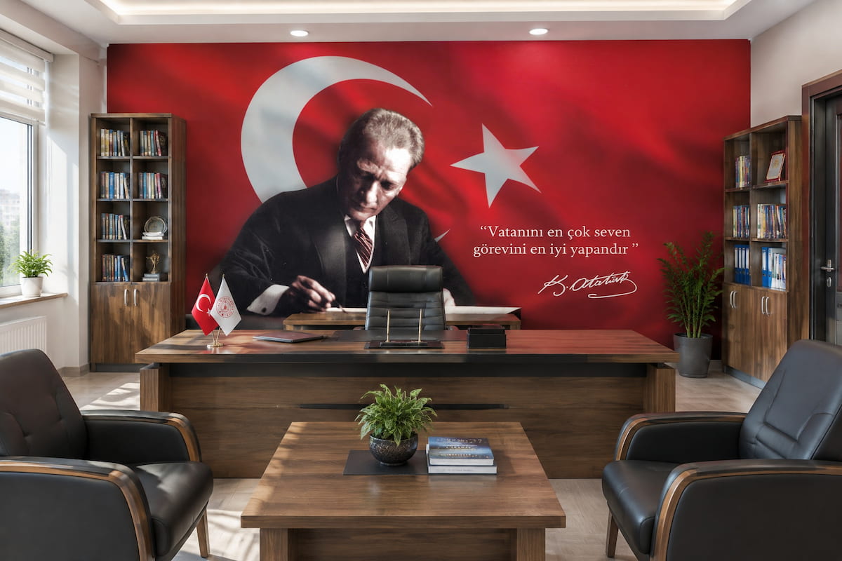 Makam odasında ahşap çalışma masası ve deri koltukların arkasında yer alan, dalgalanan Türk bayrağı ve yazı yazan Atatürk portreli "Vatanını en çok seven, görevini en iyi yapandır" sözü detaylı duvar kağıdı tasarımı.